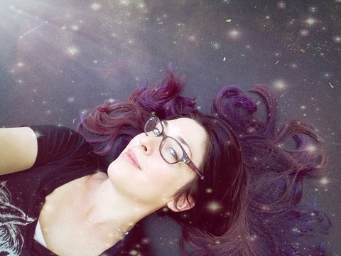 Woman with glasses rests on bed, dreaming up stellar fae aesthetic handmade jewelry ideas.