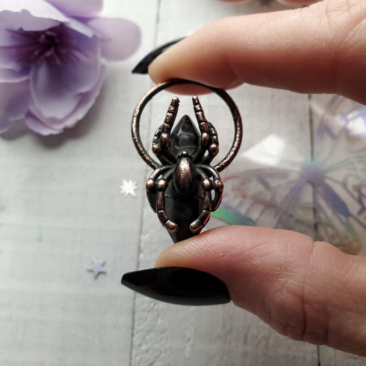 Metallic spider-shaped pendant or charm held between two fingers.